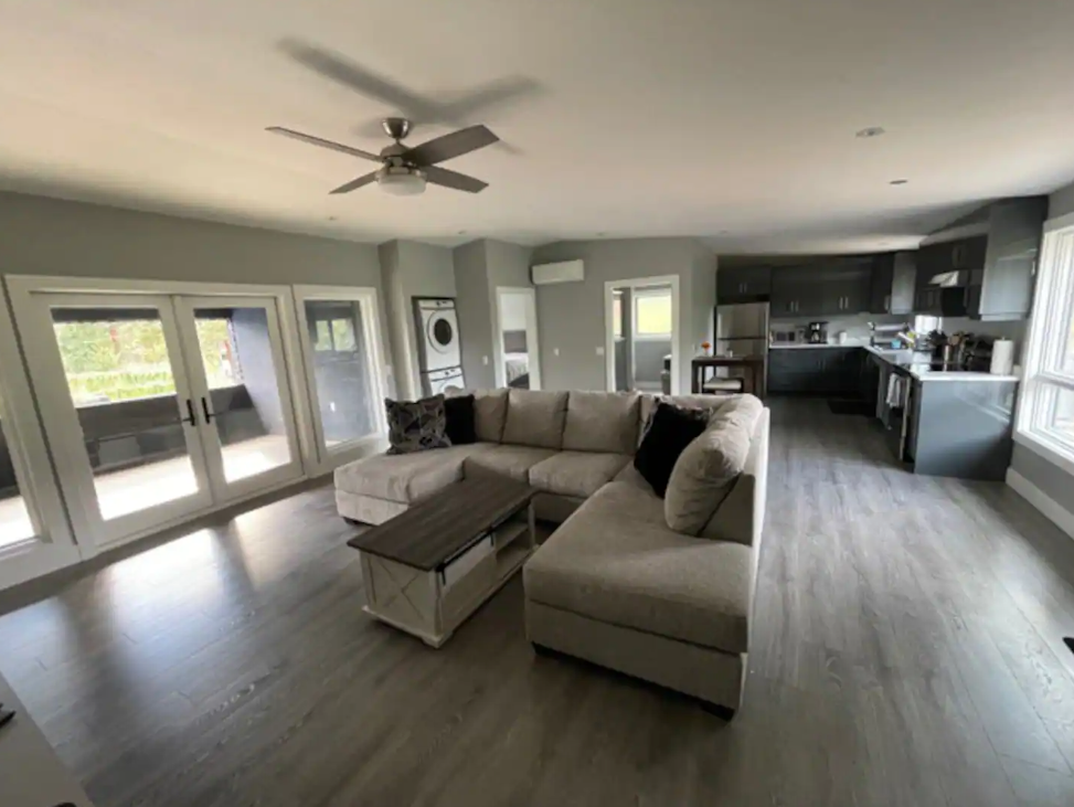 Bright living room with large sectional sofa, ceiling fan, and open kitchen area in accommodation.