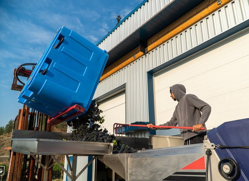 Person in hoodie using a trolley to handle a large blue container outside industrial building.